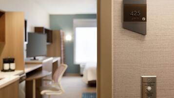 Una habitación de hotel moderna con una pantalla digital que muestra el número de habitación 425, un escritorio empotrado con una computadora, y un área de descanso con una silla.