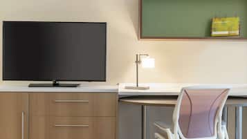Ett modernt hotellrum med en platt-TV, ett skrivbord med en lampa, en stol och en anslagstavla med en mapp.