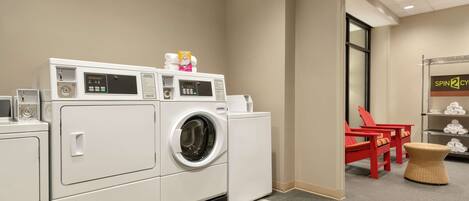 Un espace buanderie comprenant un lave-linge et un sèche-linge à chargement frontal, une petite table avec un panier, et une chaise rouge.