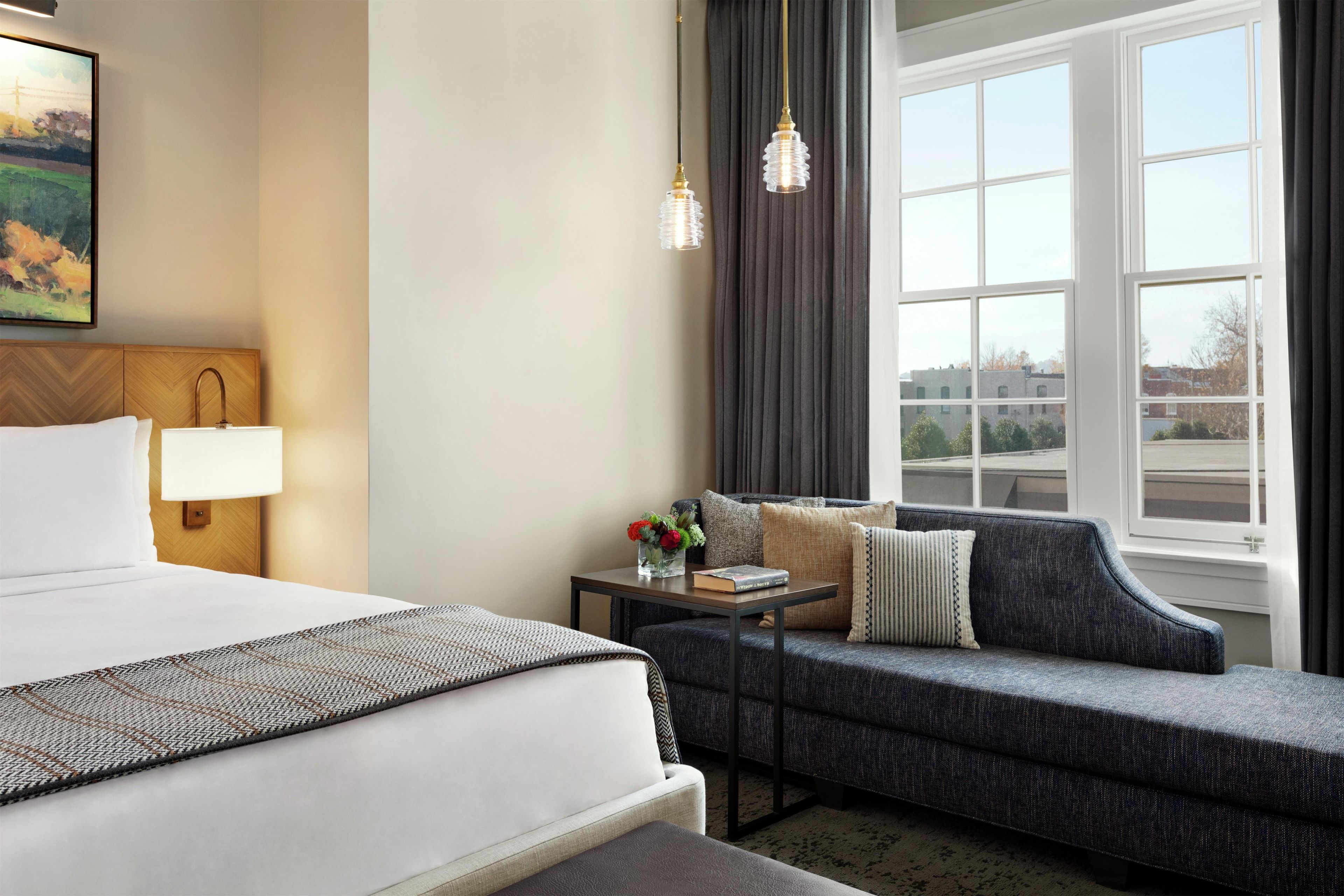A hotel room with a bed, a sofa, a nightstand, a lamp, a vase with flowers, and a window with curtains.