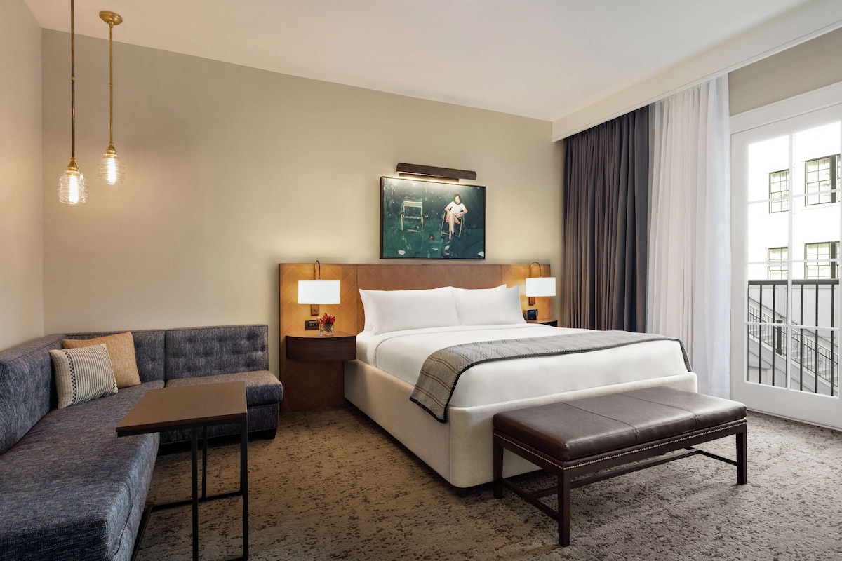 Ein Hotelzimmer mit Bett, Sofa, kleinem Tisch und einer Bank. Zwei Nachttischlampen, ein Bild an der Wand und ein Fenster mit Vorhängen sind vorhanden.