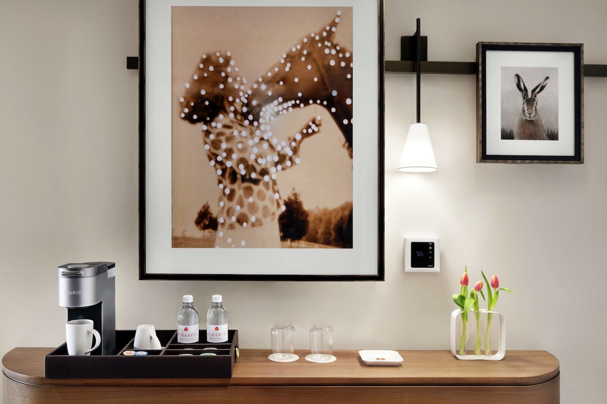 Ein Hotelzimmer mit einem gerahmten Giraffenbild, einer Kaffeemaschine, einem Tablett mit Tassen und Wasserflaschen sowie einer Vase mit Tulpen.