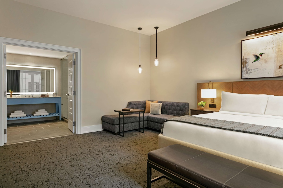 Ein Hotelzimmer mit einem großen Bett, einem grauen Ecksofa, einem kleinen Tisch, einer Bank, einem beleuchteten Spiegel und einem Wandbild.