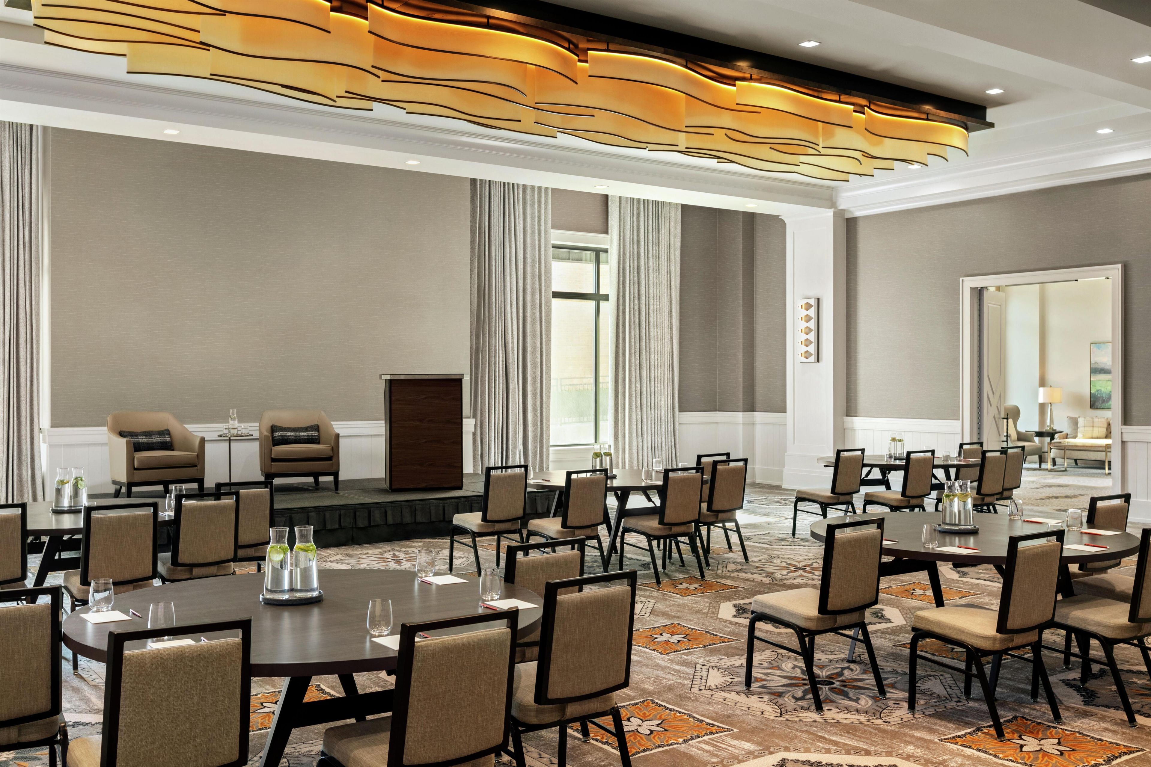 Une salle de conférence d’hôtel avec plusieurs tables et chaises, une scène et une grande fenêtre avec des rideaux.