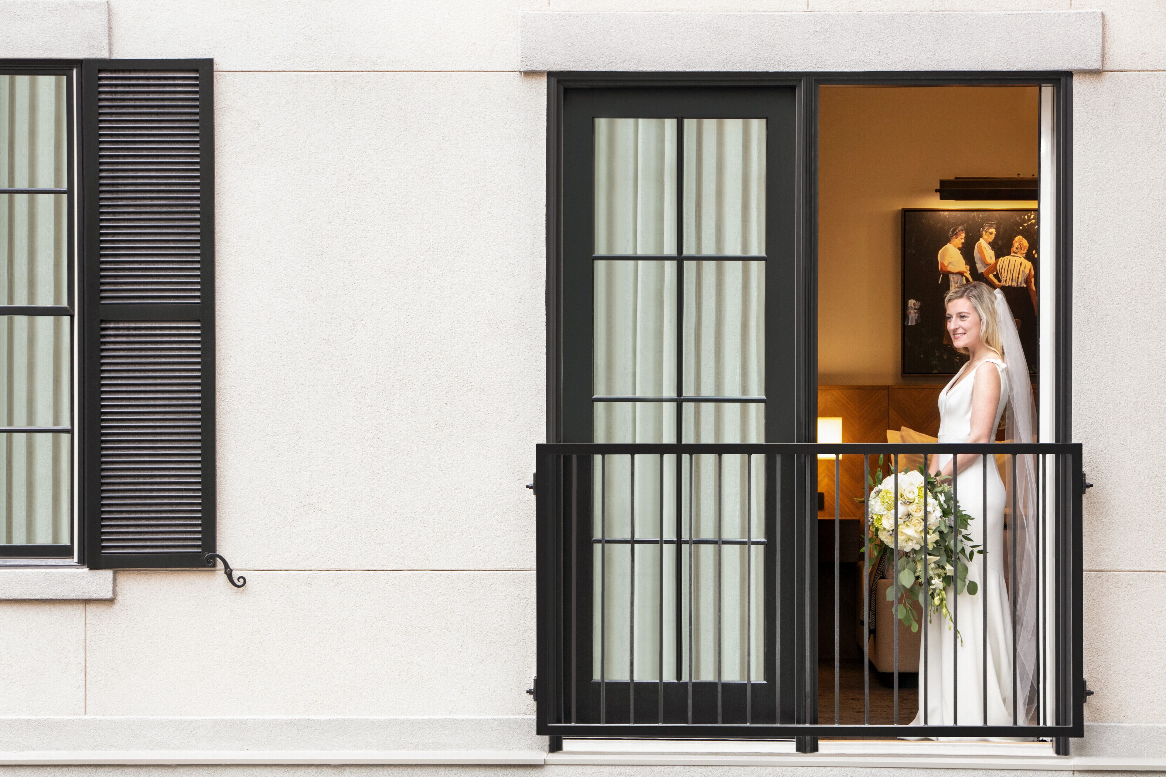 A balcony with a black railing, a window with vertical blinds, and a person holding a bouquet.