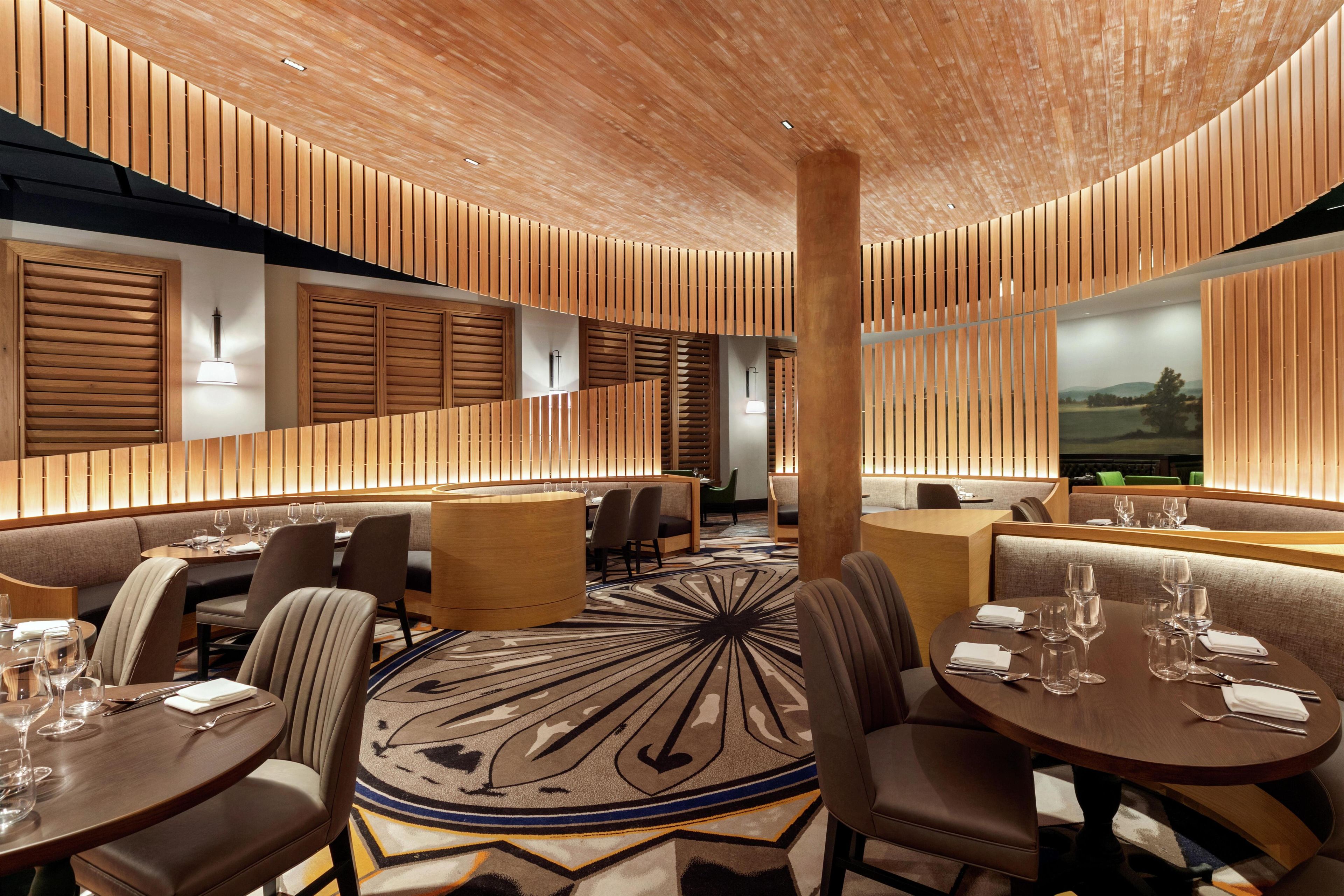 A modern restaurant with wooden paneling, round tables, and a patterned carpet.