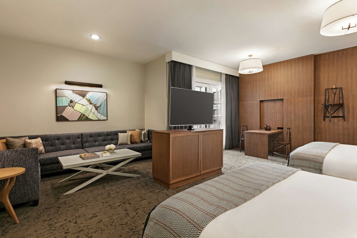 Ein modernes Hotelzimmer mit einer Couch, einem Fernseher auf einer Holzkommode, einem Schreibtisch und einem Bett.