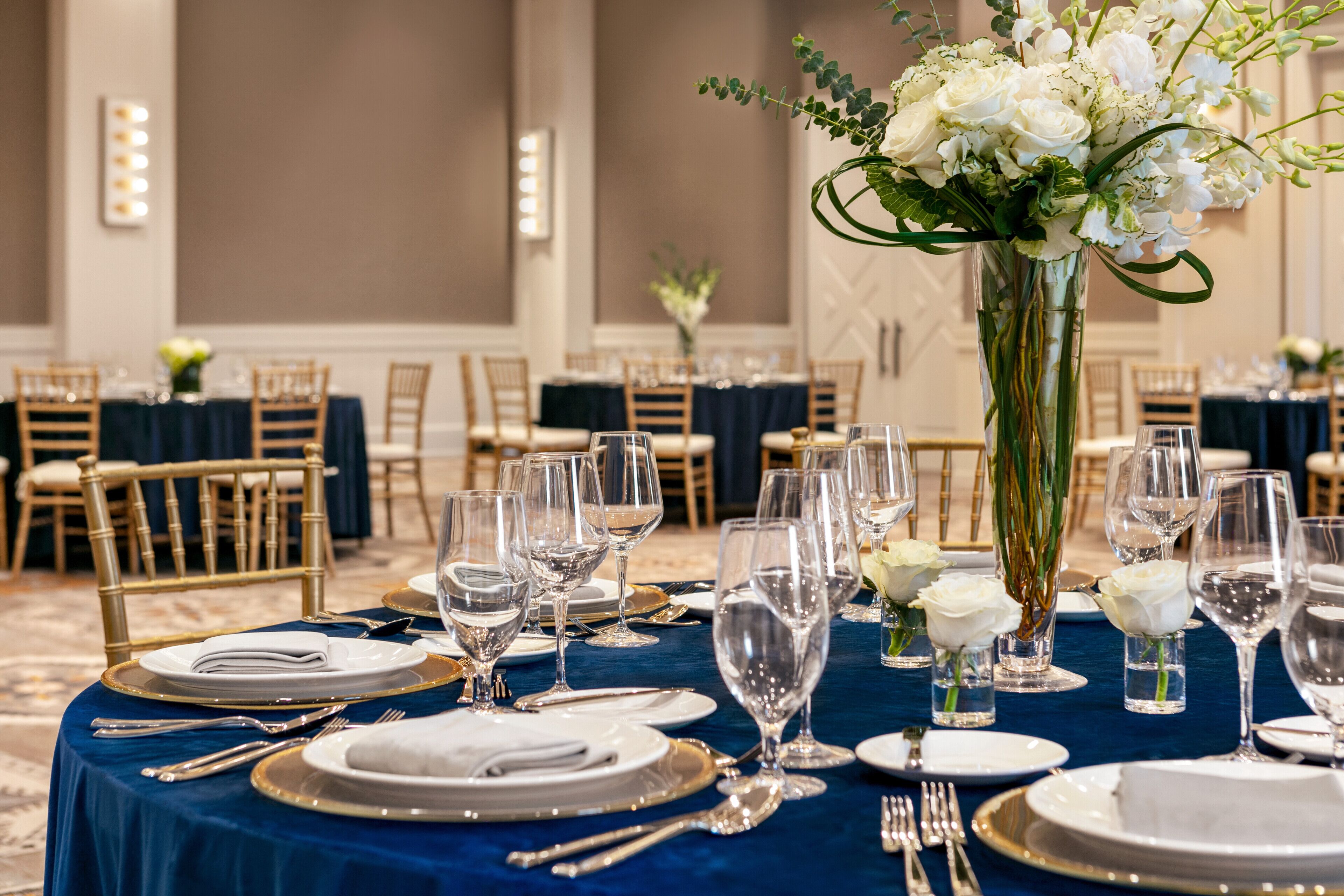 Une salle de banquet avec des tables rondes dressées pour un événement formel, agrémentée de nappes blanches, d’assiettes à bord doré et de verres en cristal.