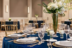 Un salón de banquetes con mesas redondas dispuestas para un evento formal, con manteles blancos, platos con ribete dorado y cristalería transparente.
