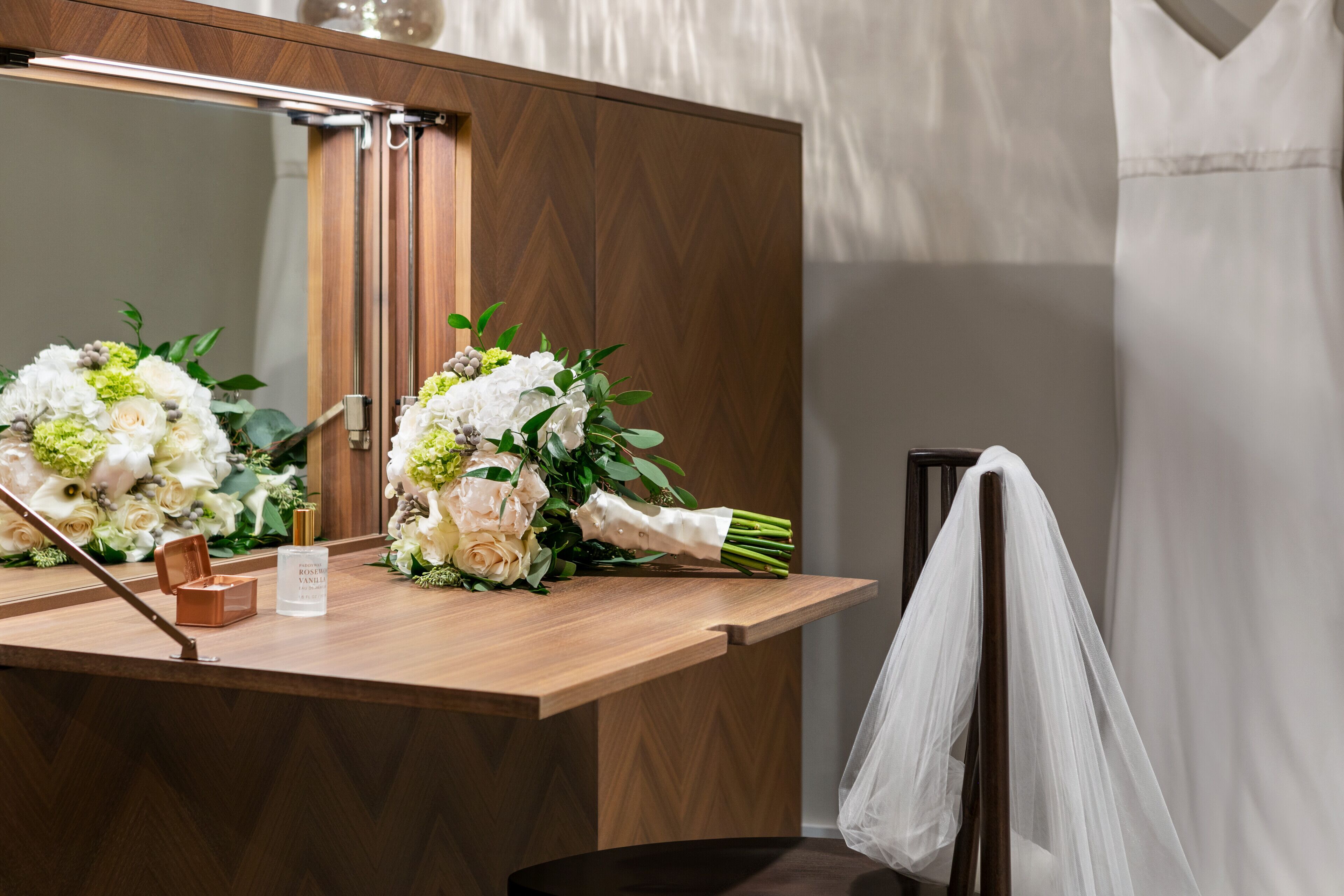 Une coiffeuse en bois avec un miroir, un bouquet de fleurs et un flacon de parfum.
