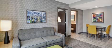 A hotel room with a grey sofa, a green ottoman, a dining area with yellow chairs, and two artworks on the walls.
