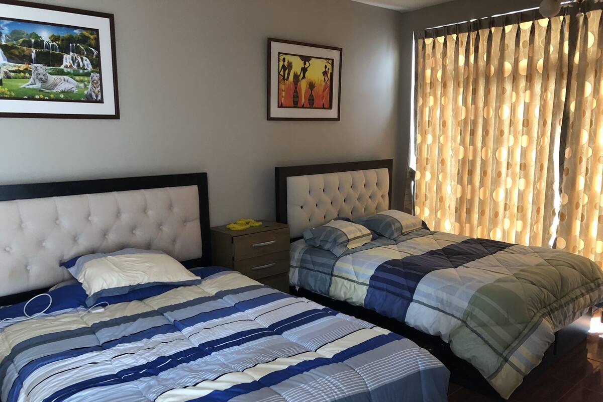 Quarto de hotel com duas camas, uma mesa de cabeceira e duas fotos emolduradas na parede.