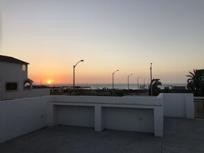 A rooftop terrace with a view of the sunset over the sea, street lamps, and a white building with arched windows. - New House by The Beach (Pisco)