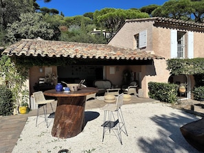 A rustic outdoor seating area with a round wooden table and wicker chairs, surrounded by a gravel patio and a building with a tiled roof and stone walls. - Villa sea view, quiet close to the center of Sainte Maxime. (Sainte-Maxime)