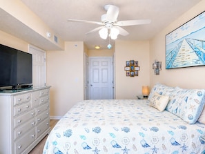 A bedroom with a bed, dresser, television, ceiling fan, and artwork on the wall.