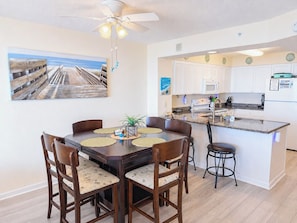 A dining area with a wooden table and chairs, a ceiling fan, and a kitchen with white cabinets and a granite countertop.