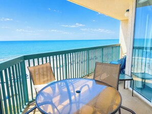 A balcony with a glass table and chairs, overlooking a beach with a clear blue sky.