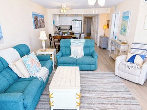 A living room with teal sofas, a white coffee table, and a dining area in the background.