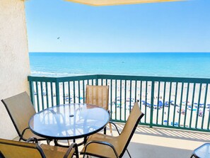 A balcony with a glass table and chairs, overlooking a beach with people and umbrellas.