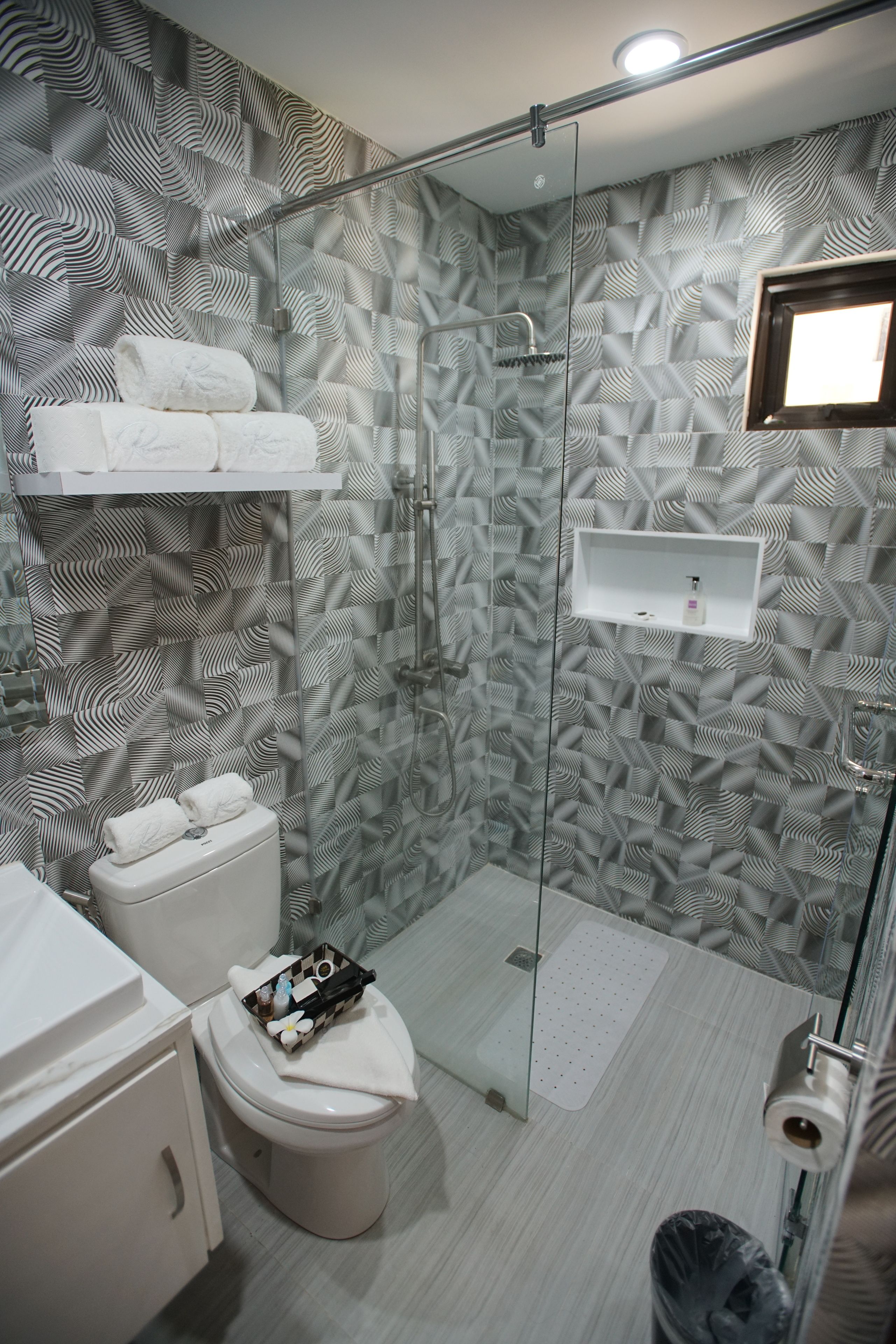 A modern bathroom with a patterned wall, a glass shower enclosure, a toilet, a sink, and a small shelf with toiletries.