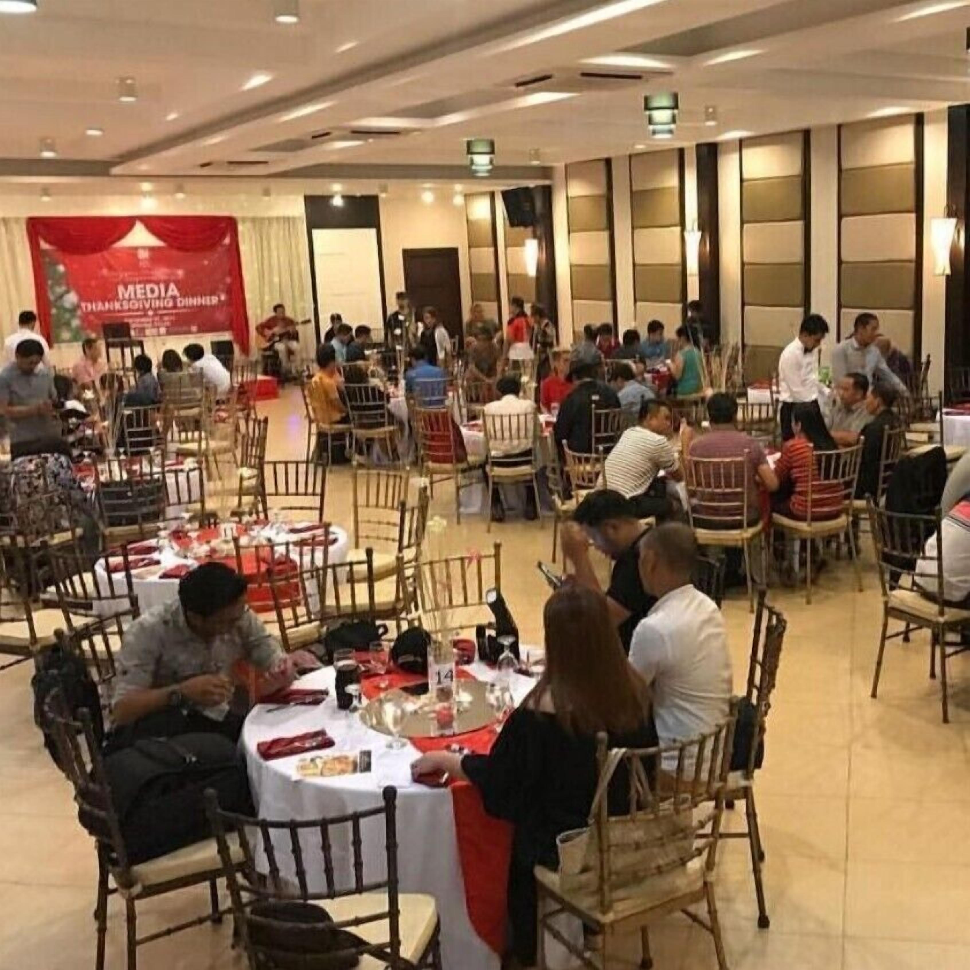 A banquet hall with round tables and chairs, a banner for a media Thanksgiving dinner, and a stage area.