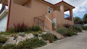 A two-story house with a balcony, a staircase leading to the entrance, and a garden with various plants and flowers. - Sunny Coco mat villa with a sea view,Cephalonia island (Kateleios)