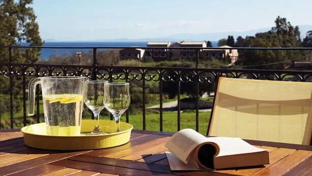 A table with a pitcher, glasses, and an open book on a balcony with a view of the sea and buildings. - Sunny Coco mat villa with a sea view,Cephalonia island (Kateleios)