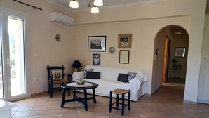 A living room with a white sofa, a round table, and a chair. There is a door with a view to the outside, an air conditioning unit on the wall, and a ceiling light fixture. - Sunny Coco mat villa with a sea view,Cephalonia island (Kateleios)