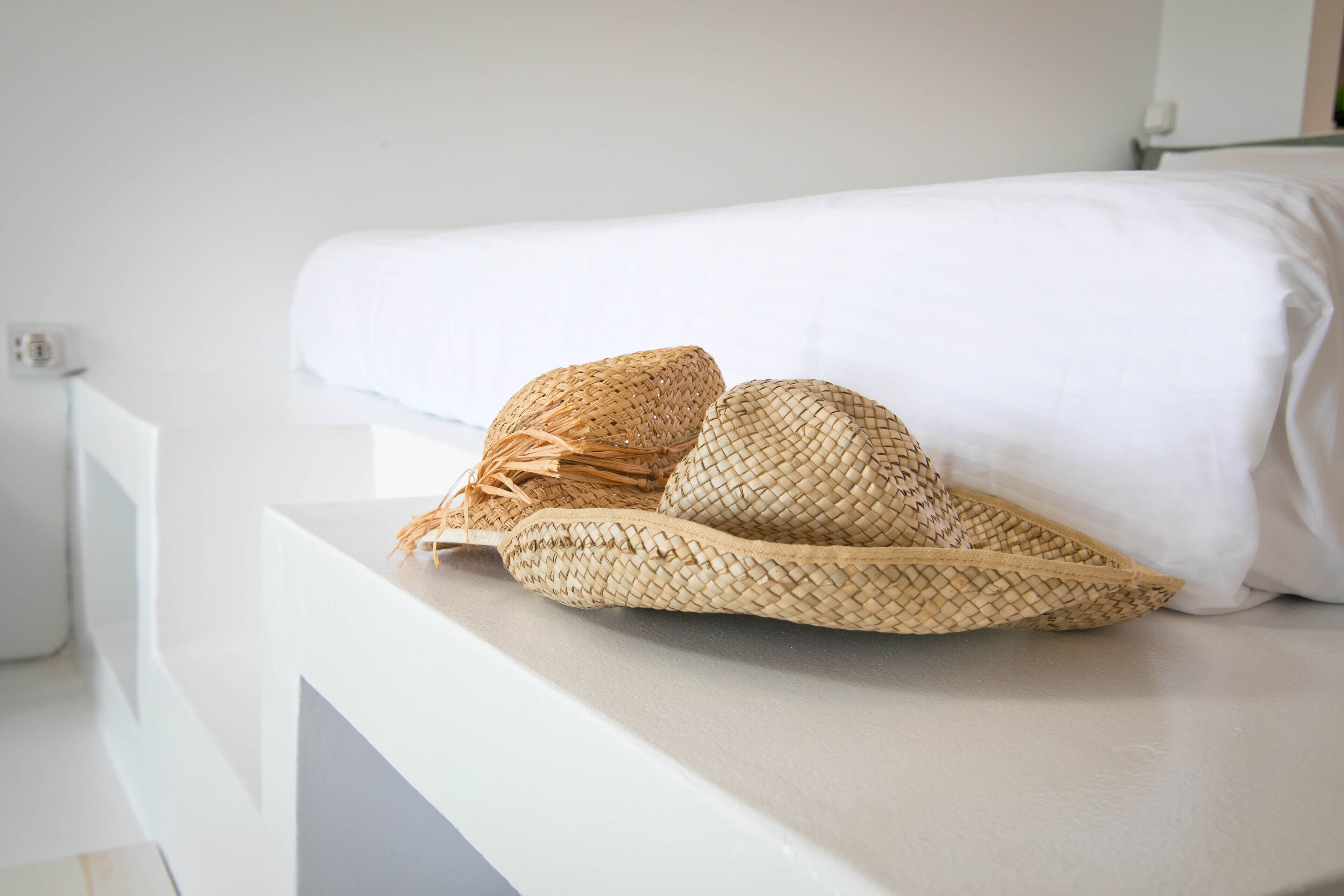 Una cama blanca con un almohadón blanco y dos sombreros tejidos sobre una mesita de noche blanca.