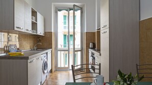 A compact kitchen with a washing machine, a dining area with a table and chairs, and a view through an open door to a balcony. - Casa Azzurra by Wonderful Italy (Genova)