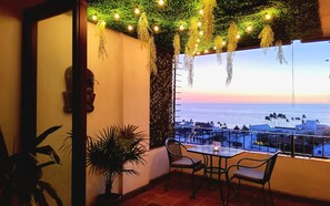 A balcony with a view of the sea, a table with chairs, hanging lights, and a decorative wall.