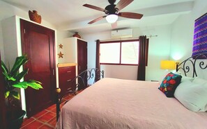 A bedroom with a bed, a ceiling fan, a window with curtains, a potted plant, and a wall-mounted air conditioner.