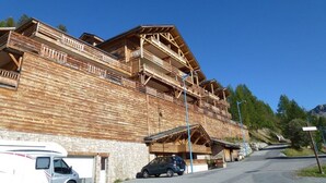Una cabaña de madera con varios balcones, zócalo de piedra y un aparcamiento con una furgoneta blanca y un coche negro.