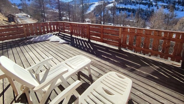 Una terraza de madera con sillas de plástico blanco y barandilla de madera que ofrece vistas a un paisaje montañoso nevado.