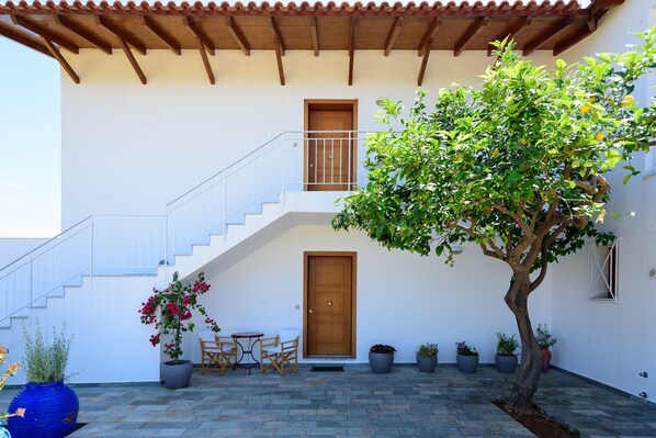 Edificio de dos pisos con una escalera de madera, un balcón y una puerta. Hay plantas en macetas y una mesa con sillas afuera.