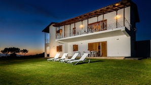 A two-story house with a balcony, a lawn with lounge chairs, and a sunset in the background.