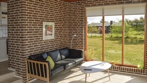 A modern living room with a brick wall, a black leather sofa, a white coffee table, and a large sliding glass door leading to a garden. - Beach front home in Sæby with WiFi (Sæby)