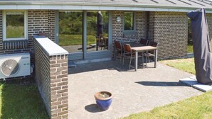 A patio area with a brick wall, a table with chairs, and a large window overlooking a garden. - Beach front home in Sæby with WiFi (Sæby)