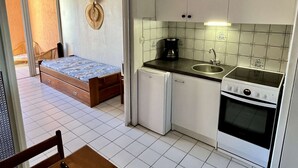 A compact kitchen with a sink, stove, and small refrigerator. A bed with a blue patterned bedspread is visible in the adjacent room. - Cosy Studio w/ Mezzanine, Parking & Pools - 400m from Beach (Argelès-sur-Mer)
