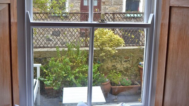 A window with a view of a stone building and a balcony with potted plants. - St Ives Cottage near beach. Sleeps 4, with Courtyard Garden + Log Burner (Saint Ives)