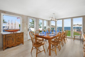 Un comedor con una mesa y sillas de madera, un modelo de barco y ventanas amplias con vista al mar.