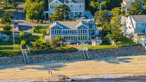 A coastal residential area with a large house featuring a balcony, a stone wall, and a beach in the foreground. - The Ultimate Nantucket Sound Beachfront Oasis! (2144) (Dennis Port)