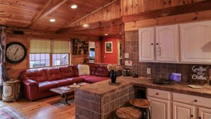 A cozy cabin interior with a wooden ceiling, a red leather sofa, a kitchen area with white cabinets, and a stone fireplace. - Stunning Lake Front Home (Glen Spey)