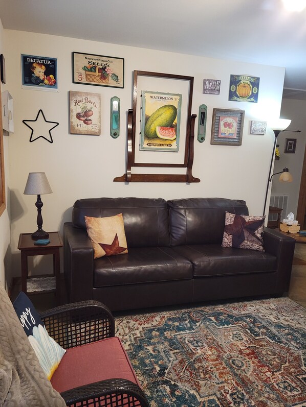 Ein Wohnzimmer mit einem dunkelbraunen Ledersofa, einem gemusterten Teppich und verschiedenen gerahmten, alten Werbeanzeigen an der Wand.