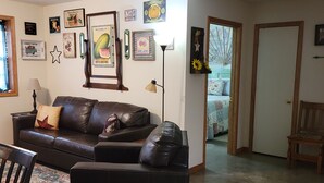 A living room with a brown leather sofa, a wooden chair, and a rug. There are wall decorations and a door leading to another room.
