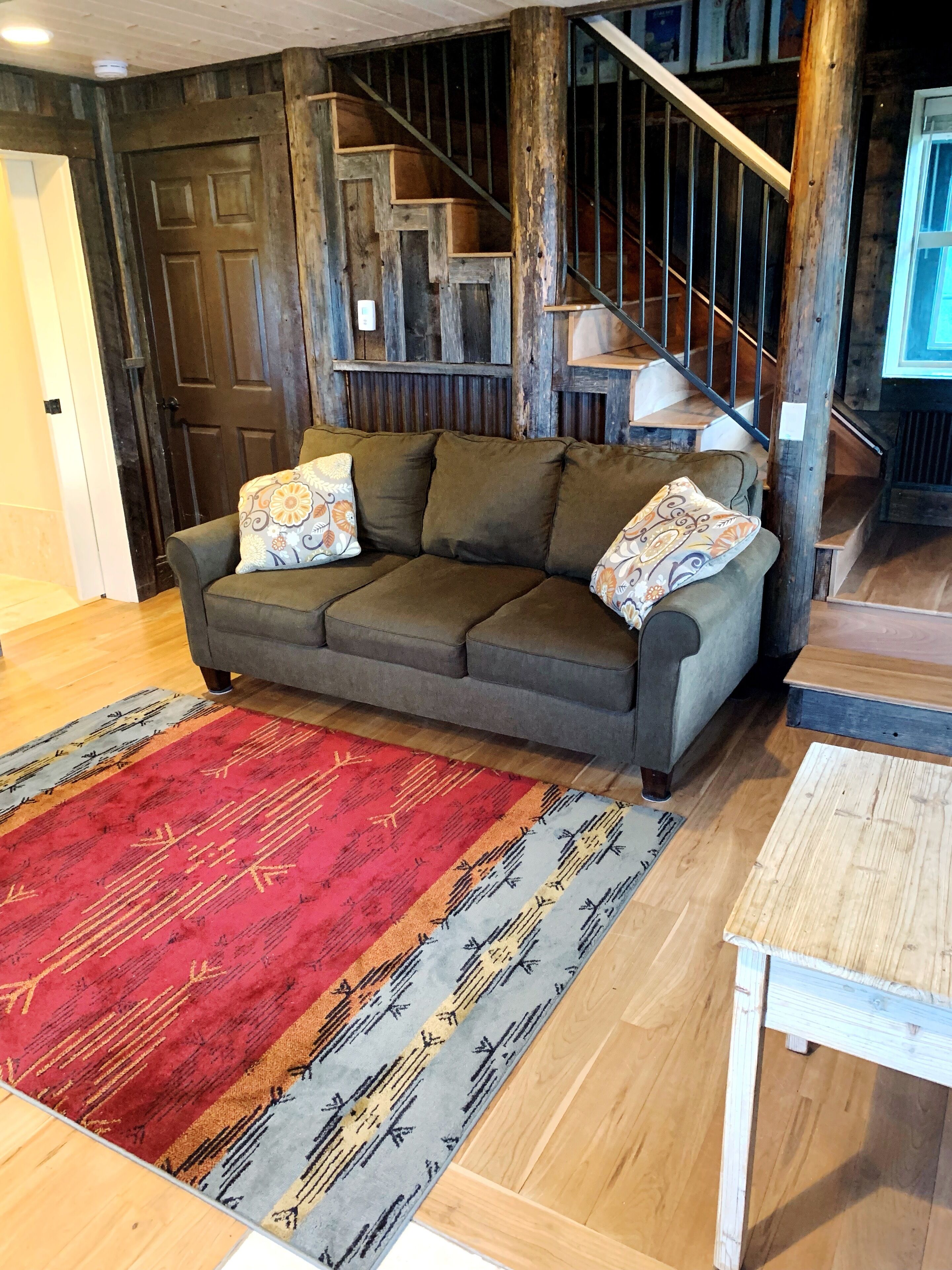A cozy living room with a grey sofa, patterned cushions, a red and grey rug, and a wooden staircase.