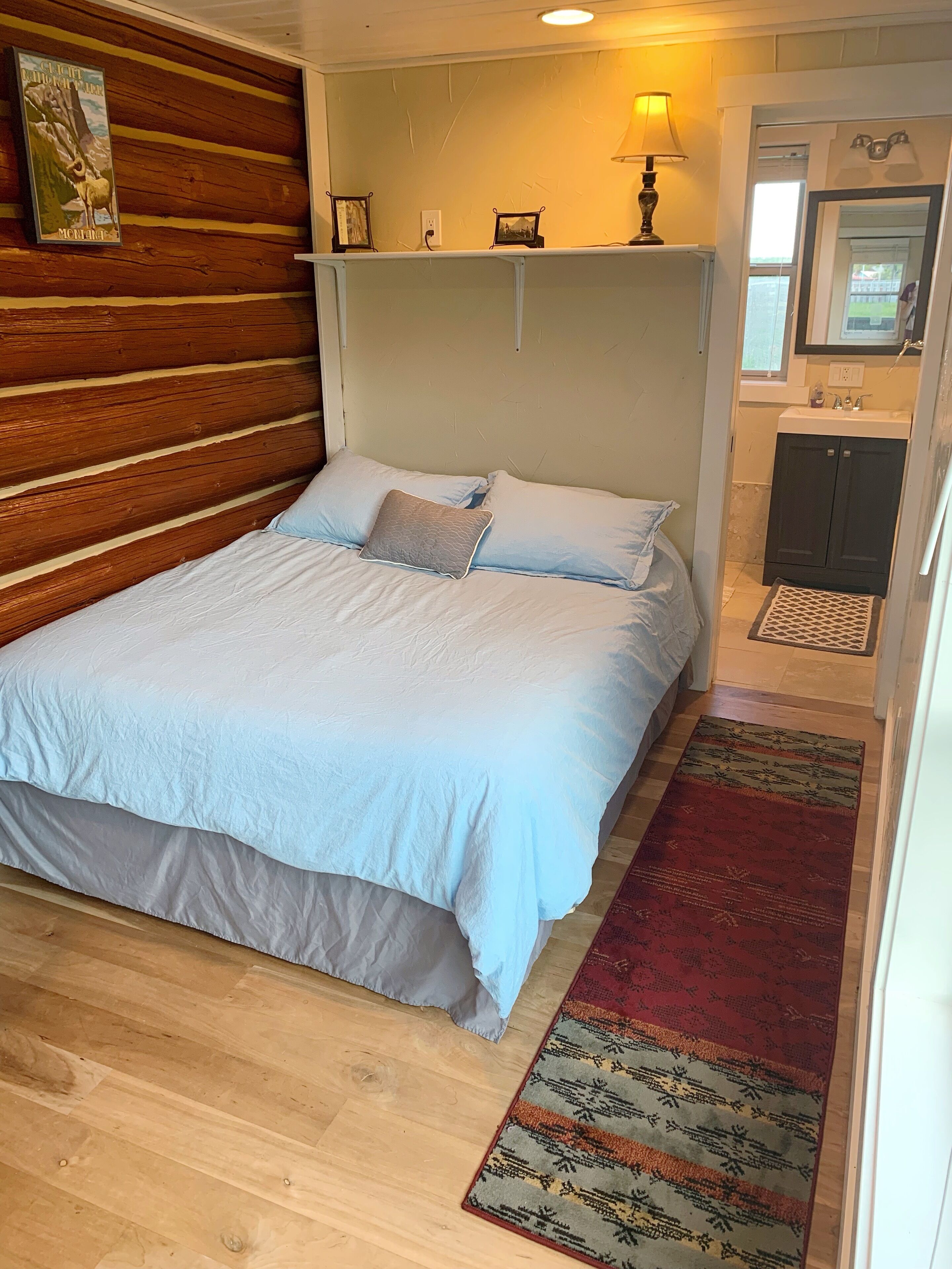 A bedroom with a wooden headboard, a bed with white bedding, a small shelf with pictures, a bedside lamp, and a bathroom with a sink and mirror.