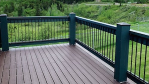 Een houten terras met een groene leuning, met uitzicht op een groen landschap met bomen en verre bergen.