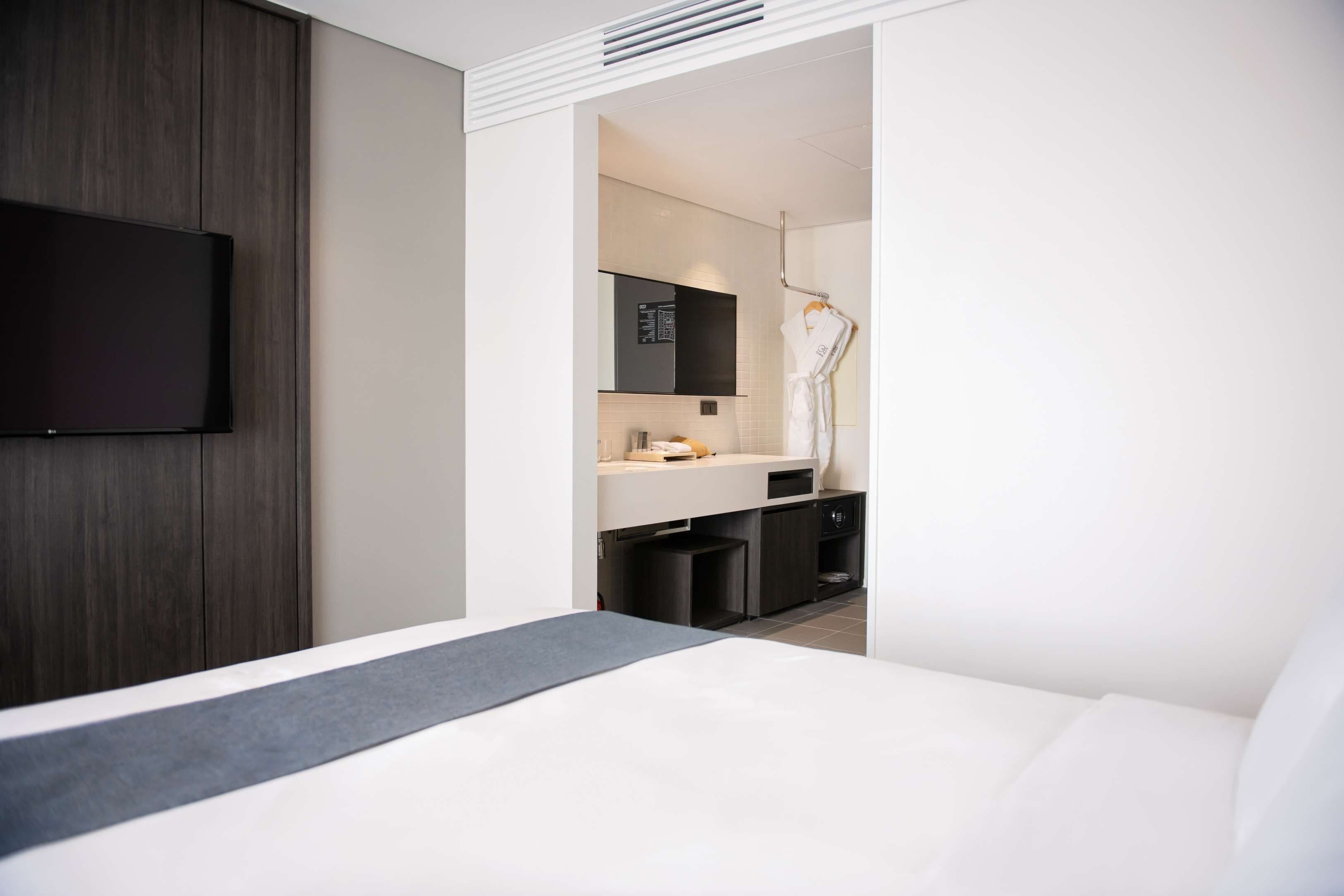 Una camera d'albergo moderna con un letto grande, una TV a schermo piatto montata a parete, un angolo cottura piccolo e un bagno visibile tramite una porta aperta.