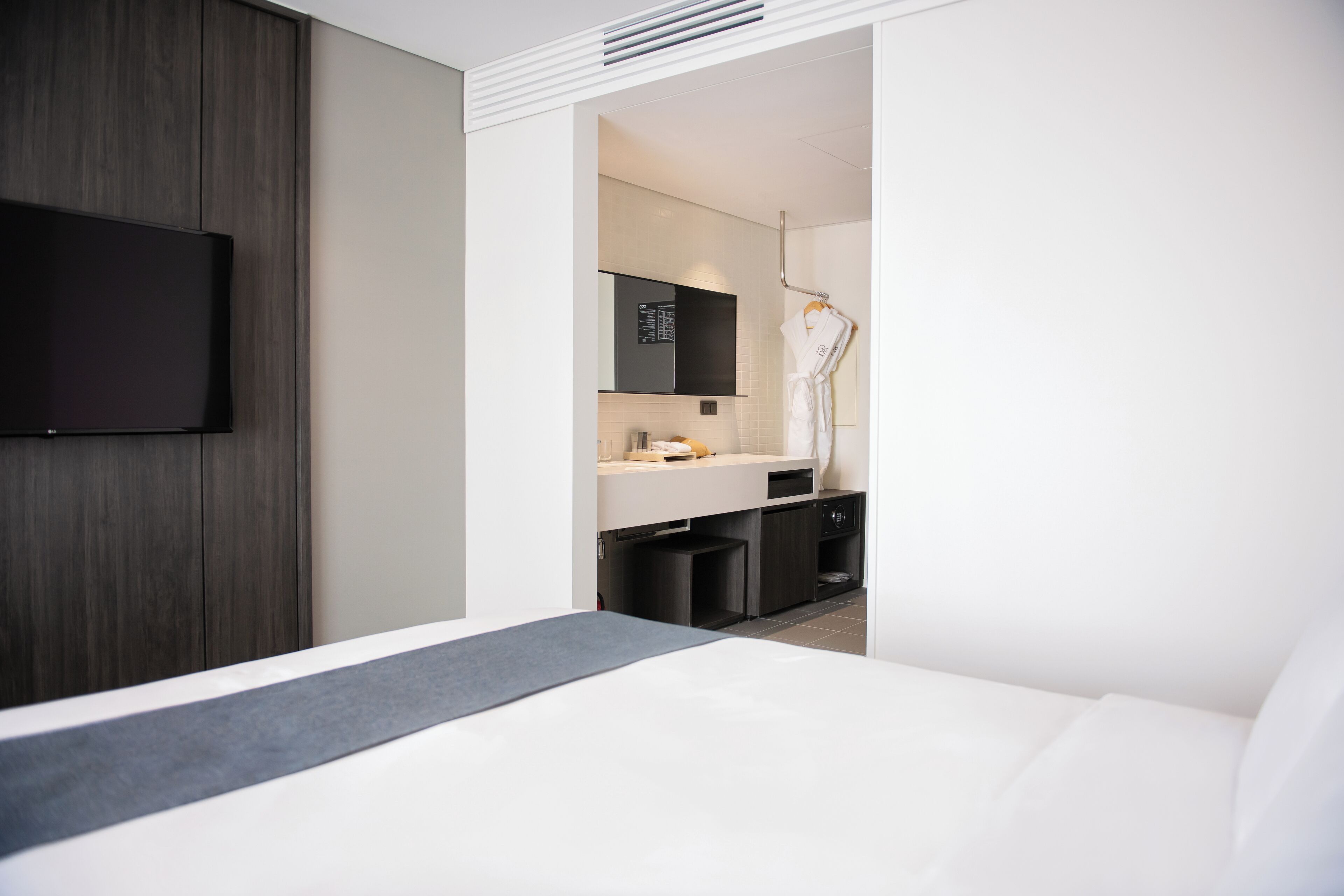 A modern hotel room with a large bed, a flat-screen TV mounted on the wall, a small kitchenette, and a bathroom visible through an open door.